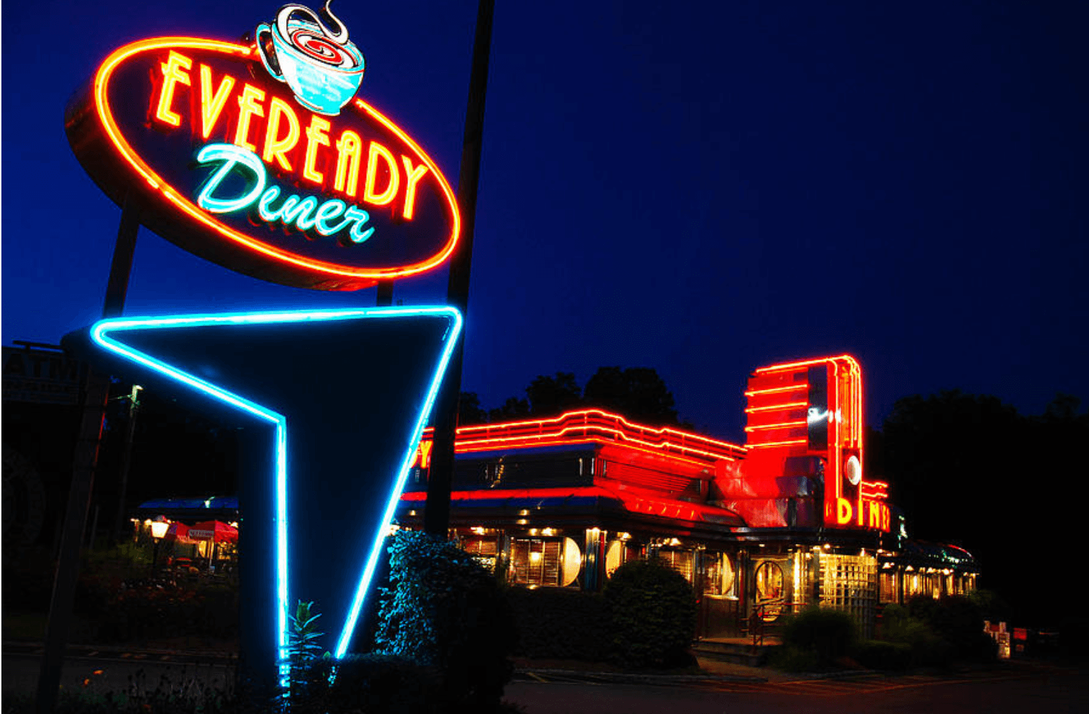 A brightly lit Eveready Diner sign glows in neon at night, as the classic diner building shines with red and orange neon lights against the deep blue sky.